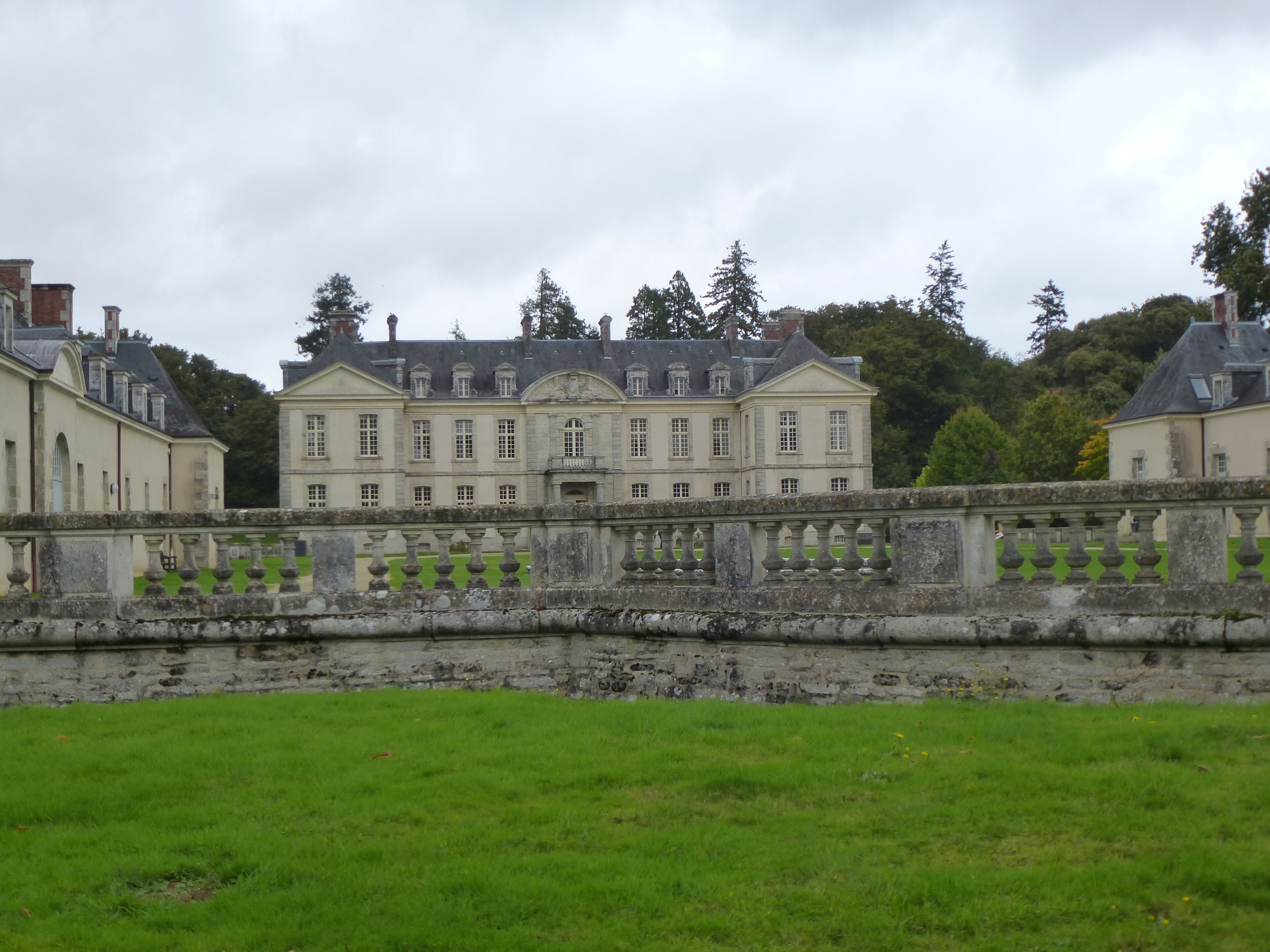 A large, historic building with a symmetrical facade, surrounded by a stone wall and lush green lawn.