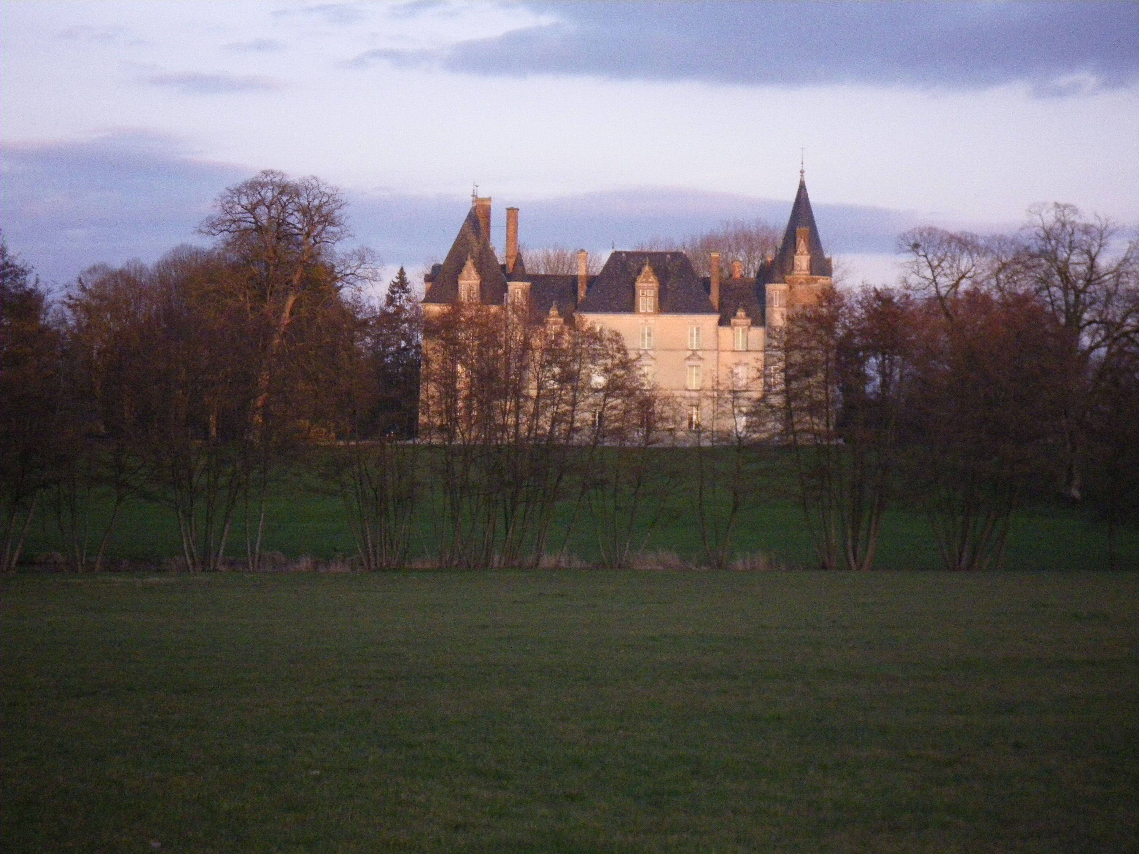 A large, historic building with multiple towers and chimneys, surrounded by trees and a grassy field.