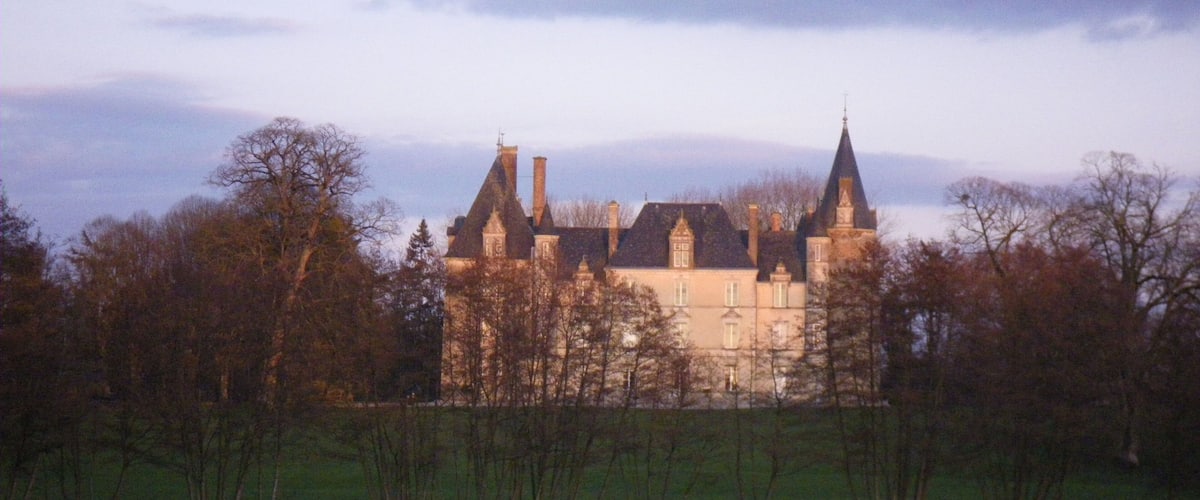A large, historic building with multiple towers and chimneys, surrounded by trees and a grassy field.