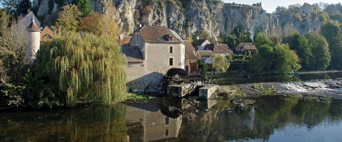 A historic castle on a cliff overlooking a river, with traditional houses and a weeping willow in the foreground.