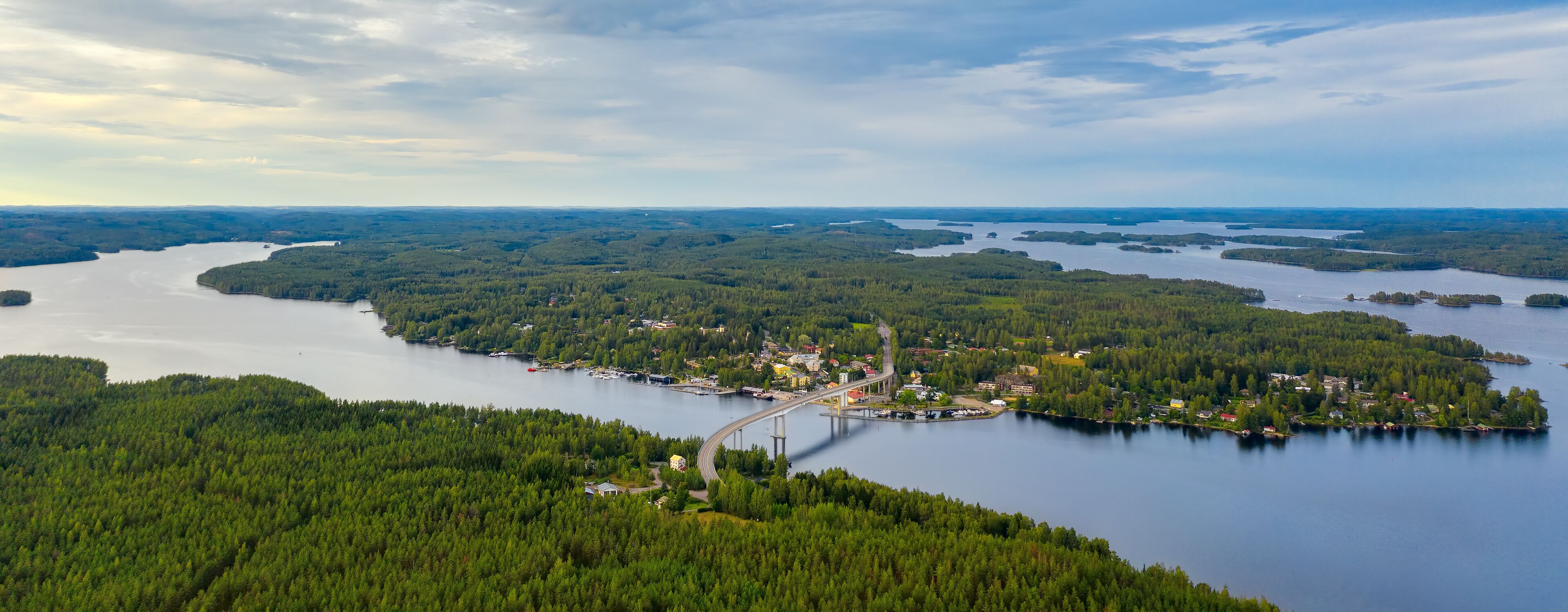 Aerial view on the bridge over the lake and trees in the forest on the shore. Blue lakes, islands and green forests from above on a cloudy summer day. Lake landscape in Finland, Puumala.