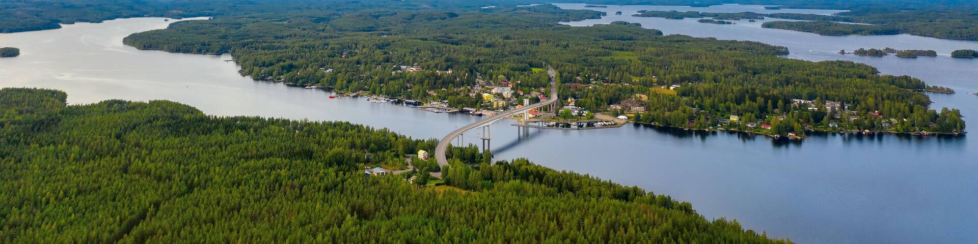 Aerial view on the bridge over the lake and trees in the forest on the shore. Blue lakes, islands and green forests from above on a cloudy summer day. Lake landscape in Finland, Puumala.