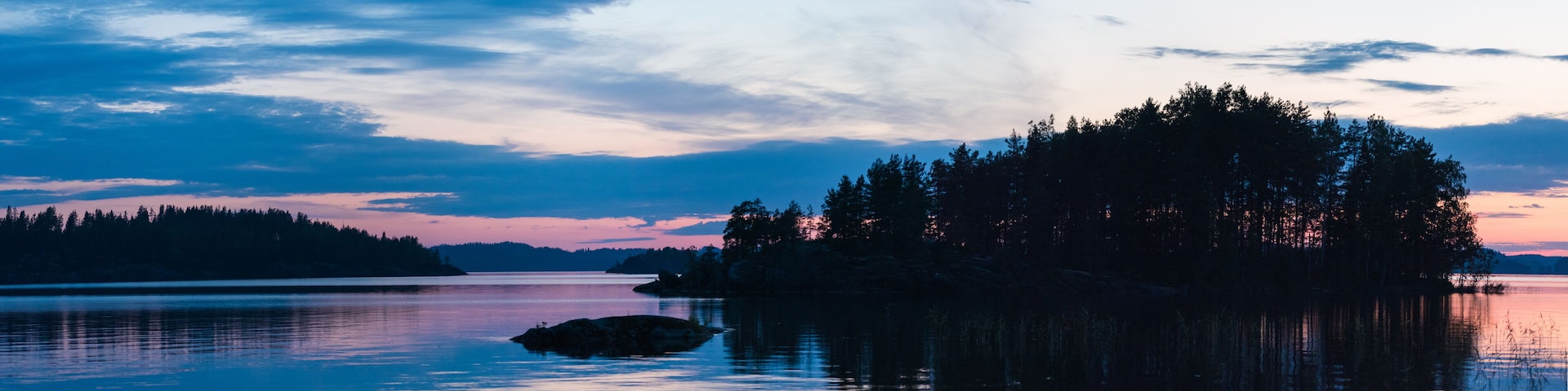 A calm lake with a small island covered in trees and a large rock in the foreground.
