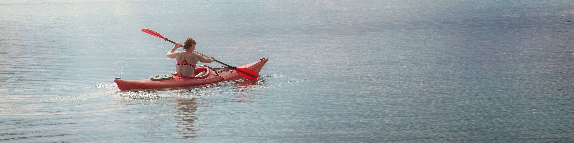 A kayaker in a red kayak on calm water with a sailboat in the distance and a rocky shoreline with trees.