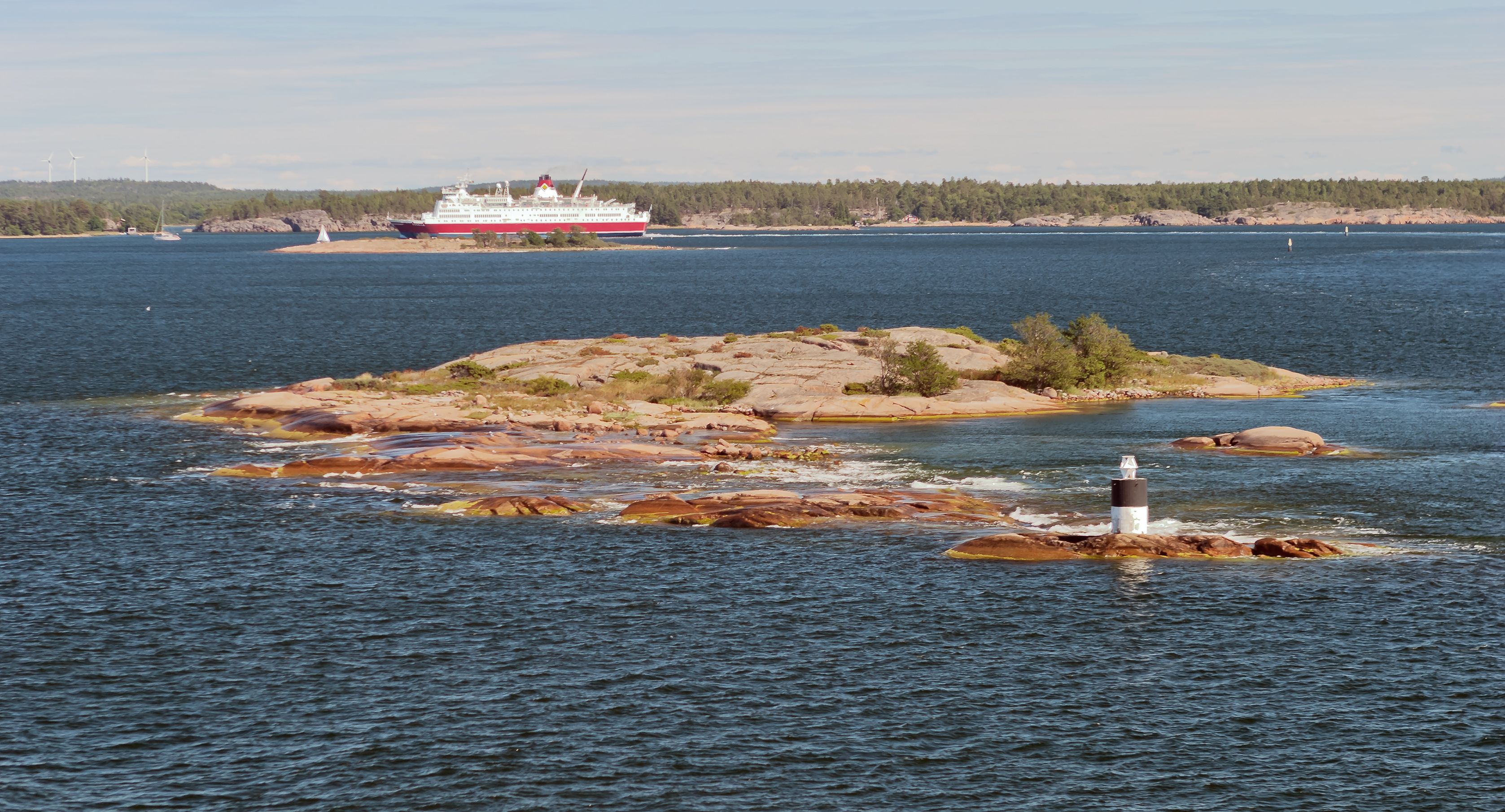 A large cruise ship in the distance, a rocky island with a lighthouse in the foreground, and a forested shoreline.