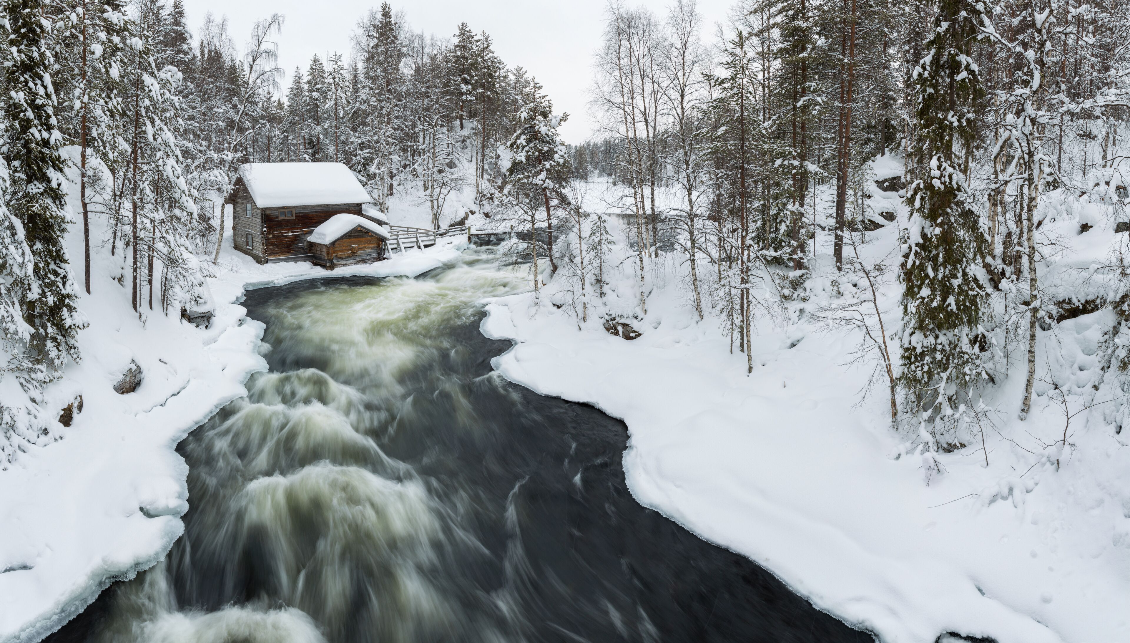 A snow-covered river flowing through a forest with a wooden cabin on the left.