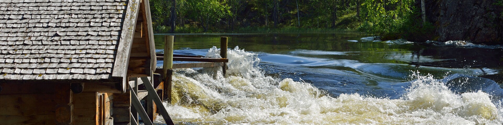 Una estructura de madera con techo de paja junto a un río de aguas rápidas, rodeada por un bosque denso.