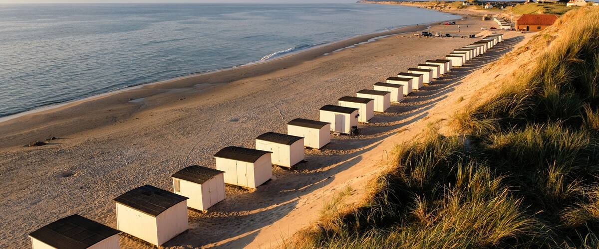 A beach with white beach huts lined up on the sand, a clear sky, and distant buildings.