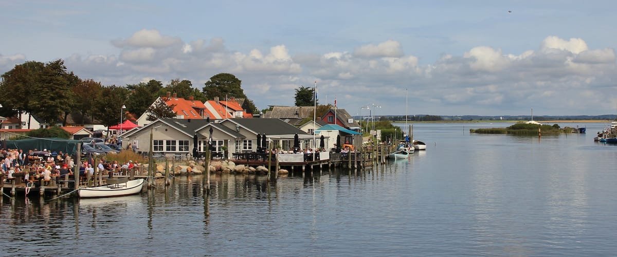 A riverside town with boats docked, a pier, and buildings with red roofs.