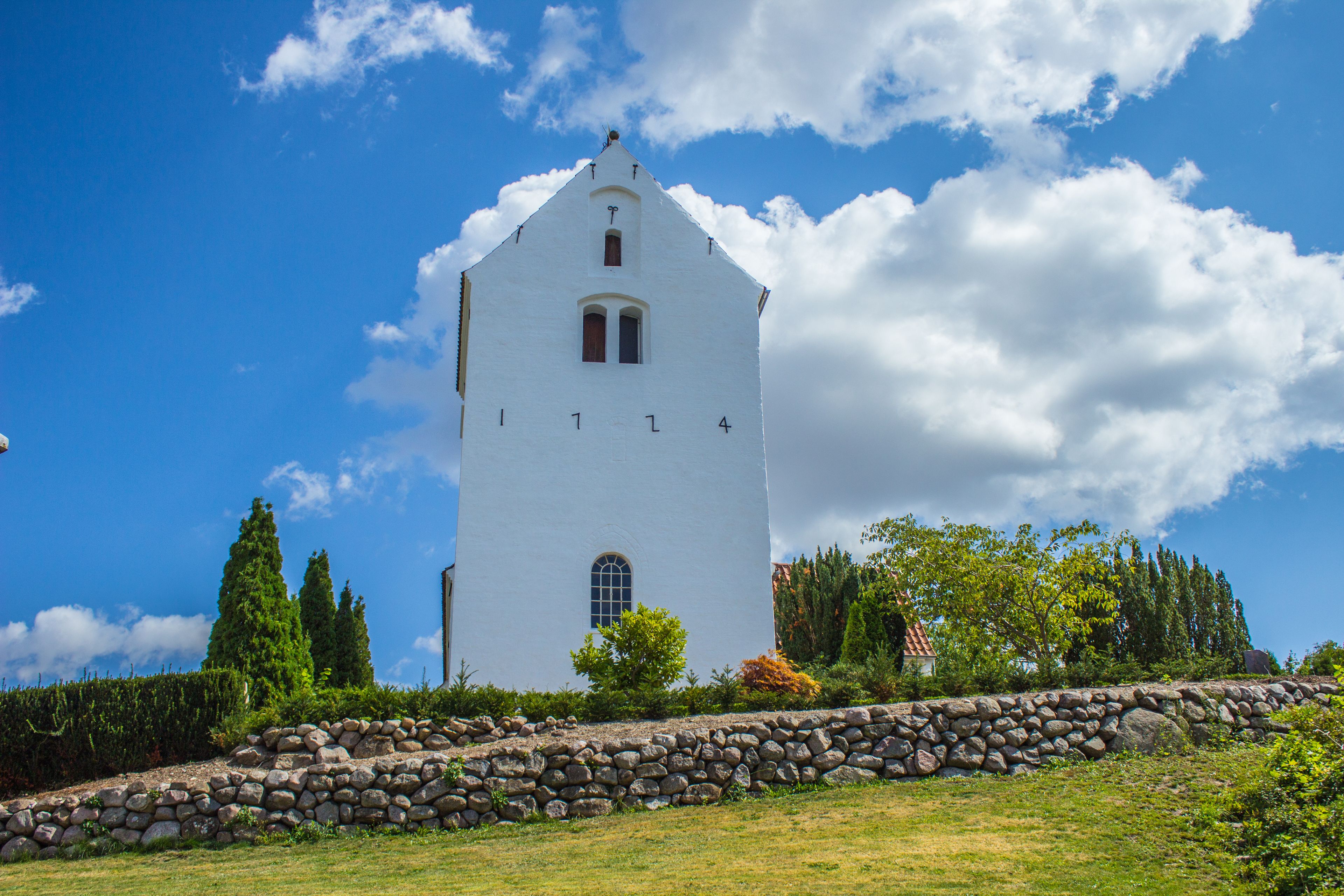 A white church with a bell tower, surrounded by trees and a stone wall.