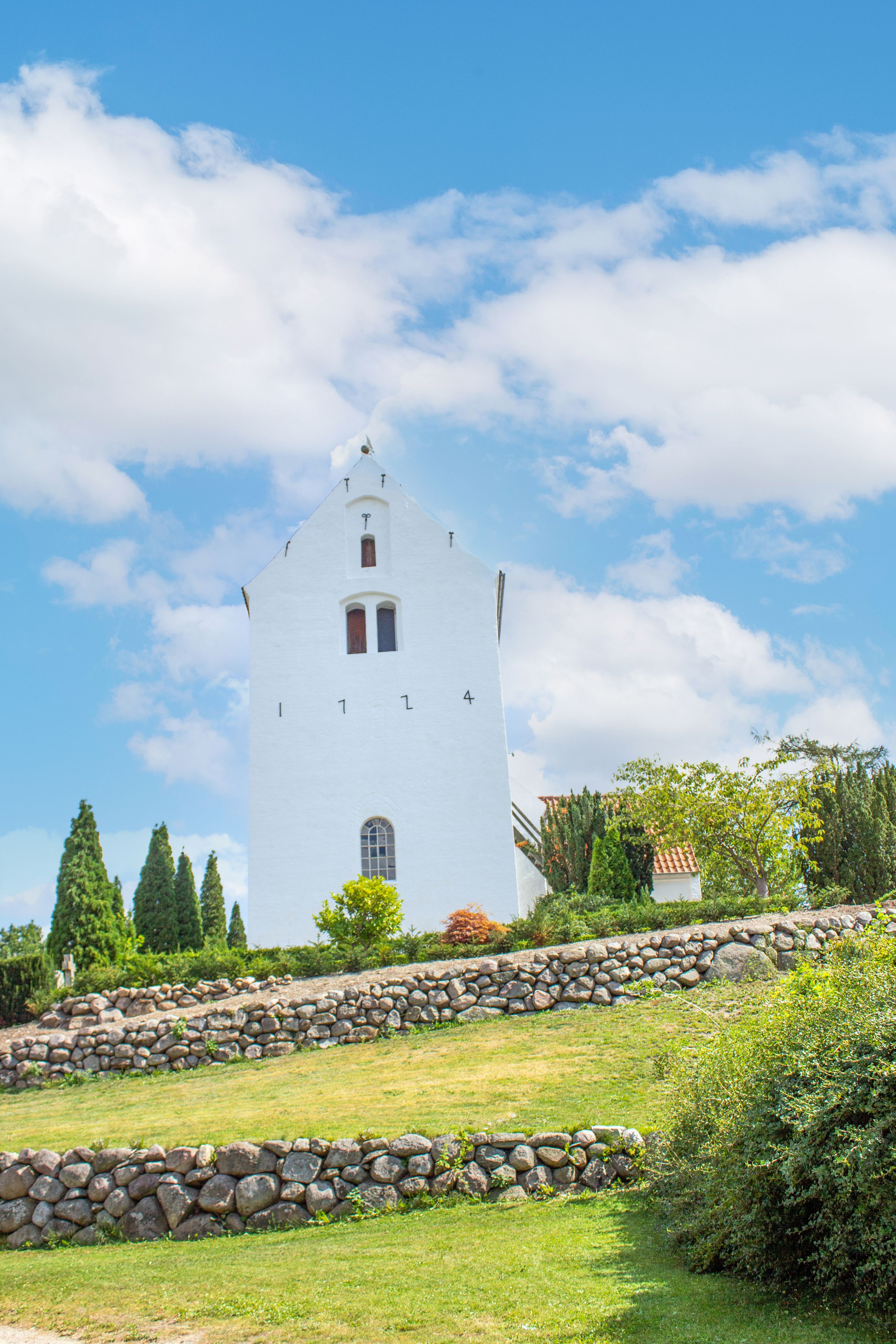 A white church with a bell tower, surrounded by a stone wall and greenery.