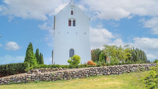 Et hvidt tårn med spids tag, omgivet af grønt og en stenvæg.