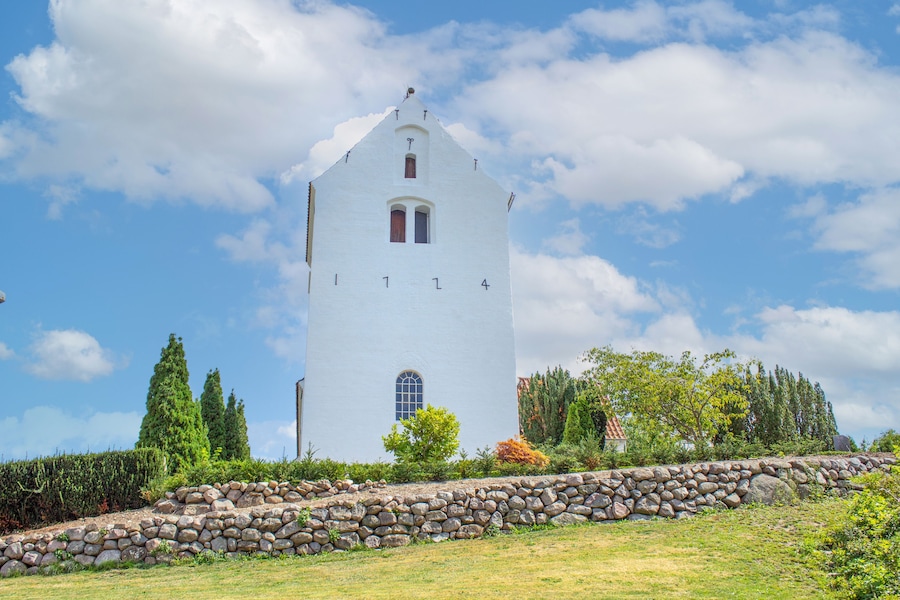 Una torre bianca con tetto a punta, circondata da vegetazione e un muro di pietra.