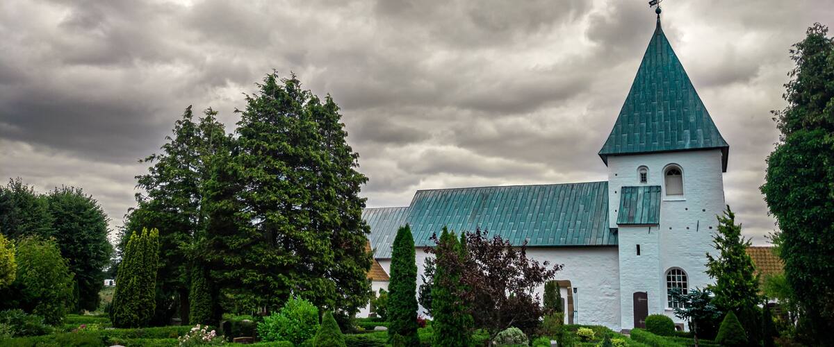 A church with a green roof and white walls, surrounded by well-manicured gardens and trees.