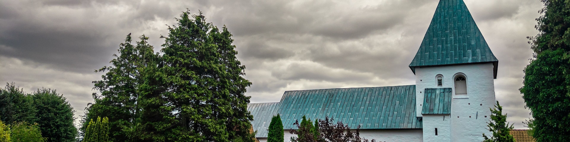 A church with a green roof and white walls, surrounded by well-manicured gardens and trees.