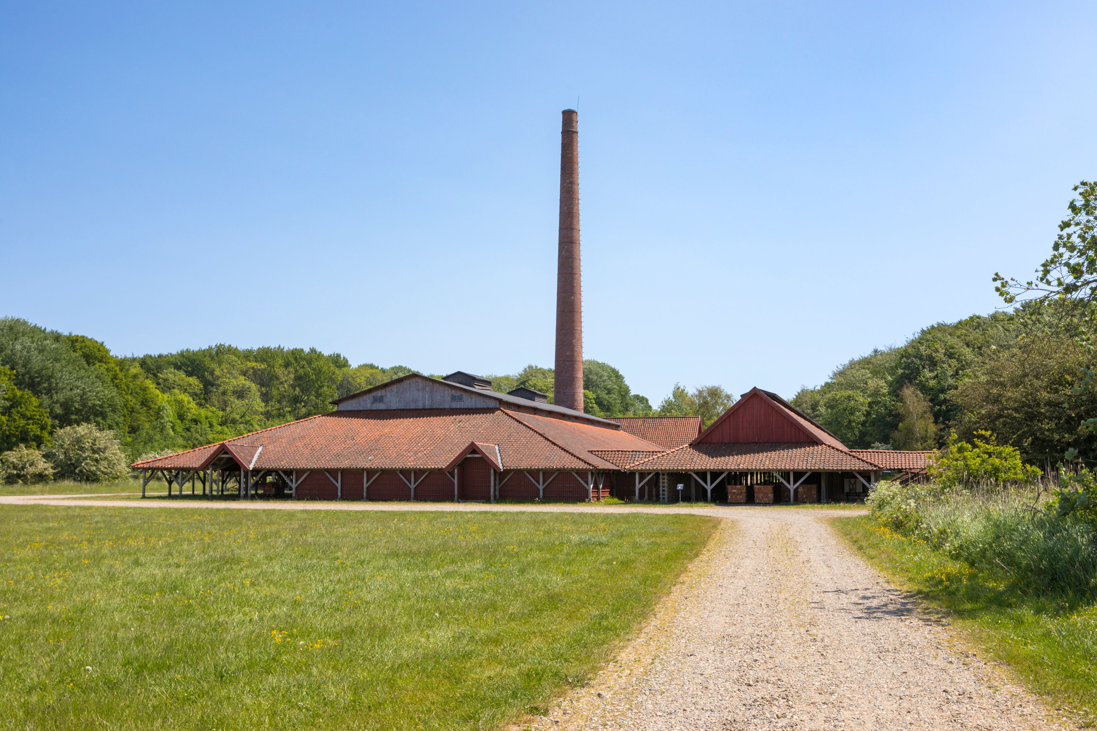A rural landscape with a large red barn, a tall chimney, and a gravel path leading to the barn.