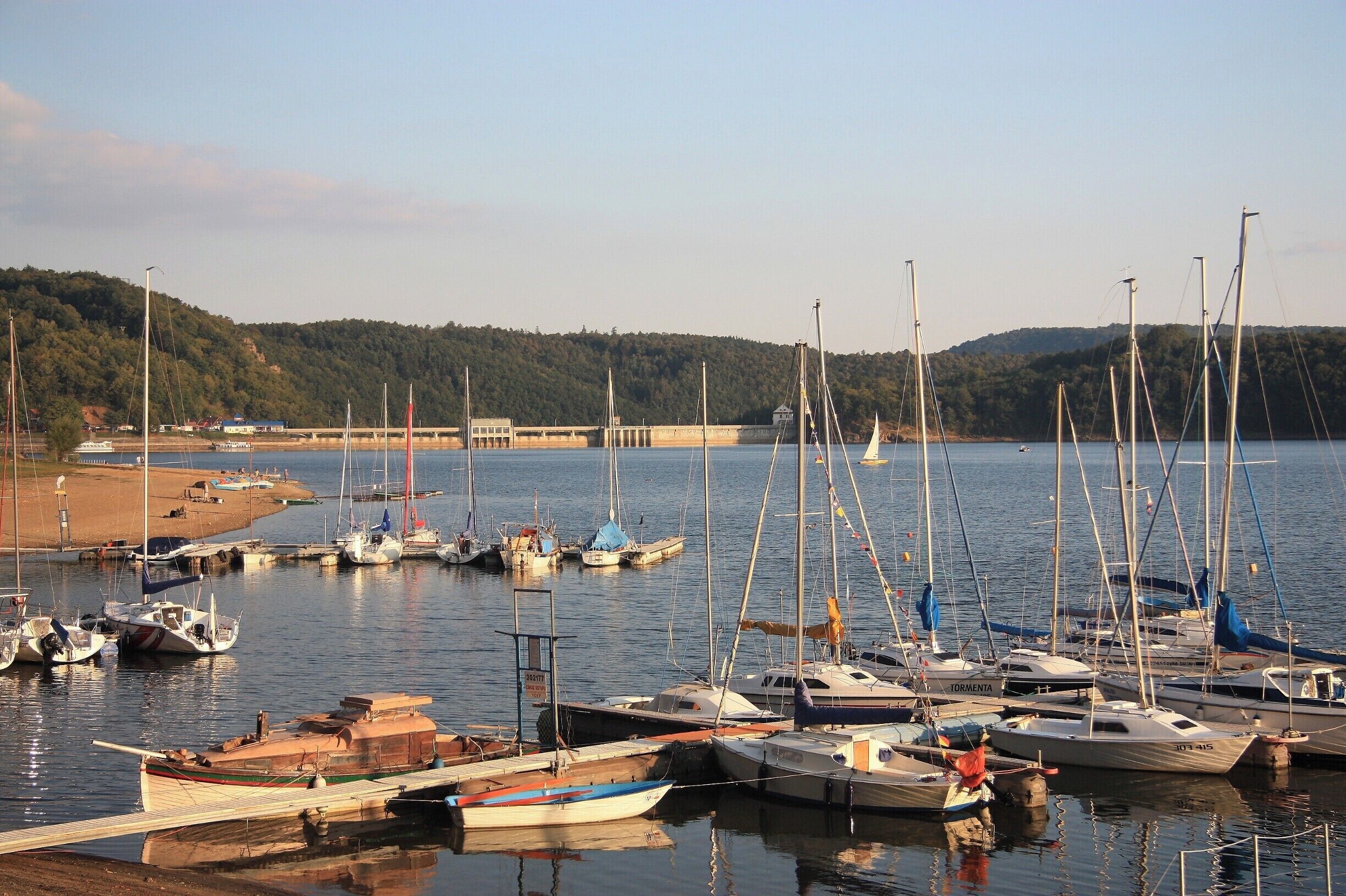 A marina with various boats docked, a sandy beach, and a forested hill in the background.