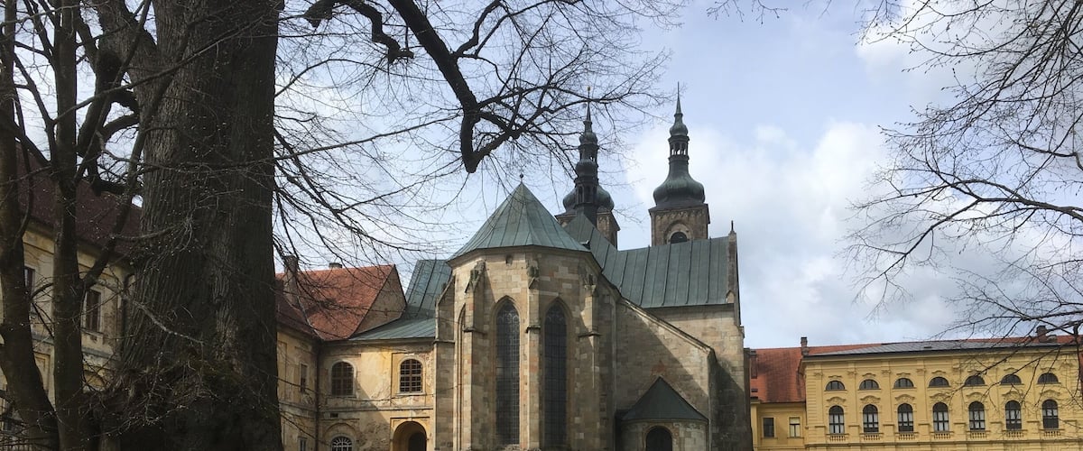 A historic church with a green roof and two spires, surrounded by trees and a grassy area with a bench.