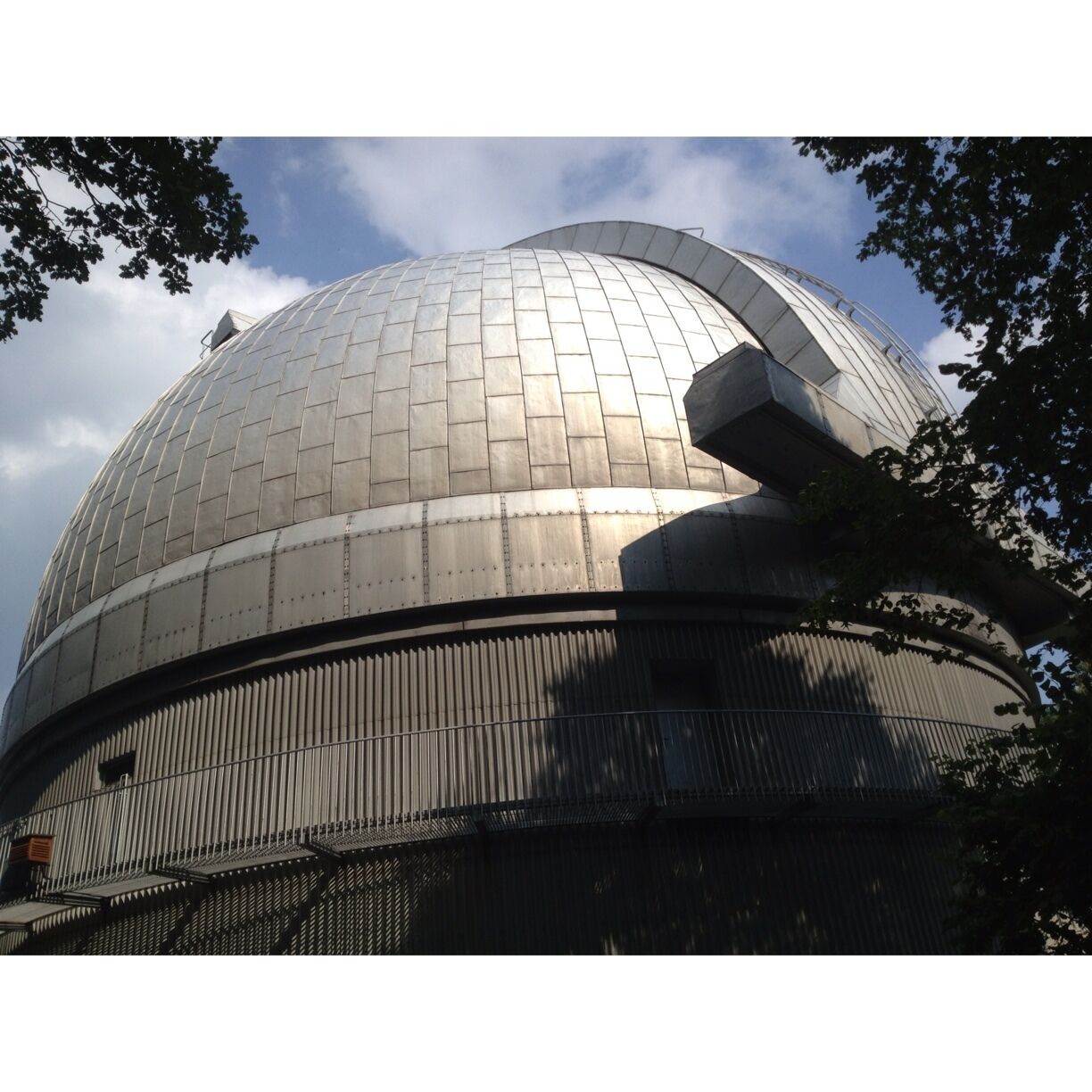 A large metallic dome structure with a grid pattern, surrounded by trees and a clear sky.