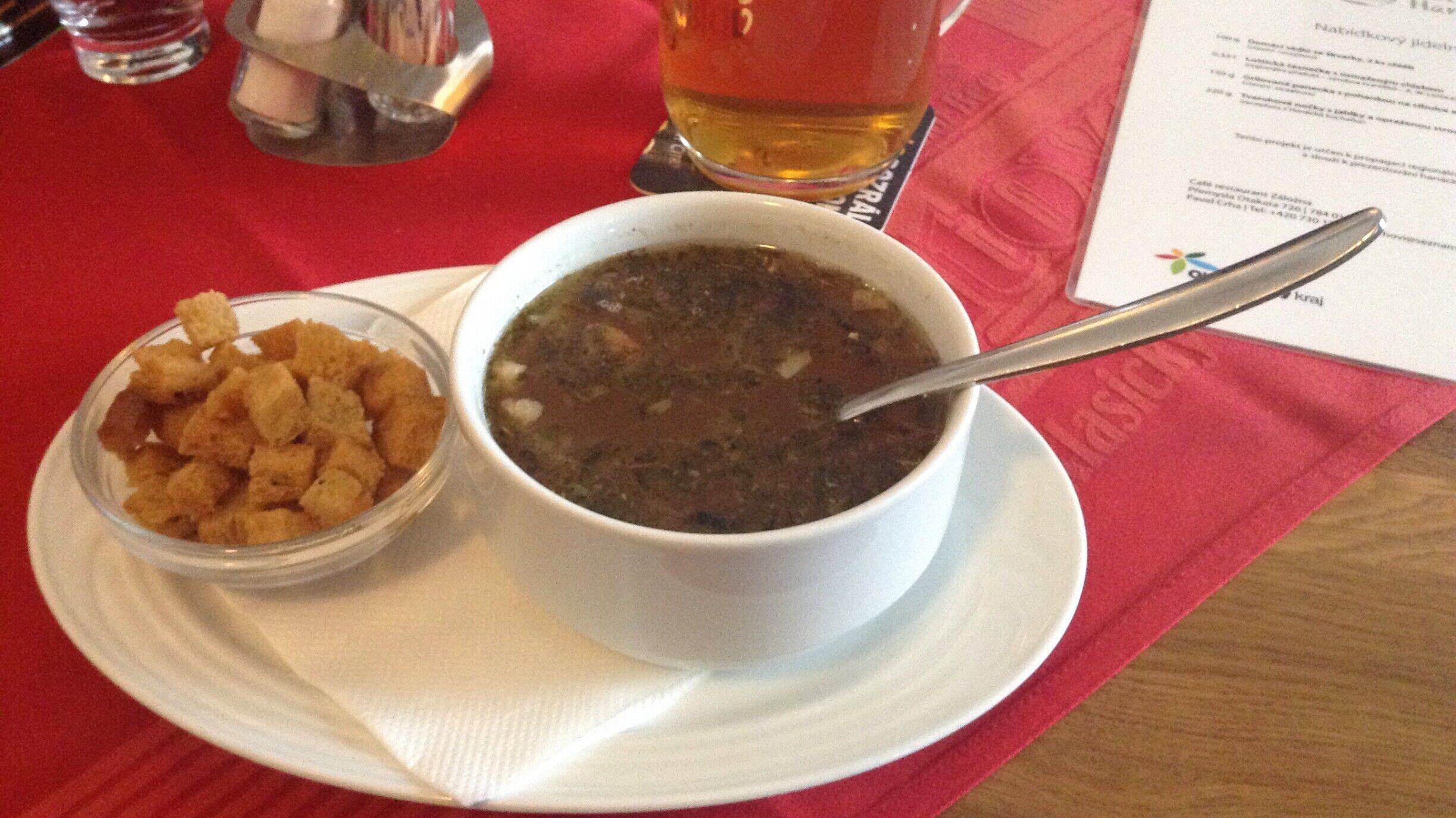 A bowl of soup with a spoon, a small bowl of croutons, and a glass of beer on a red tablecloth.