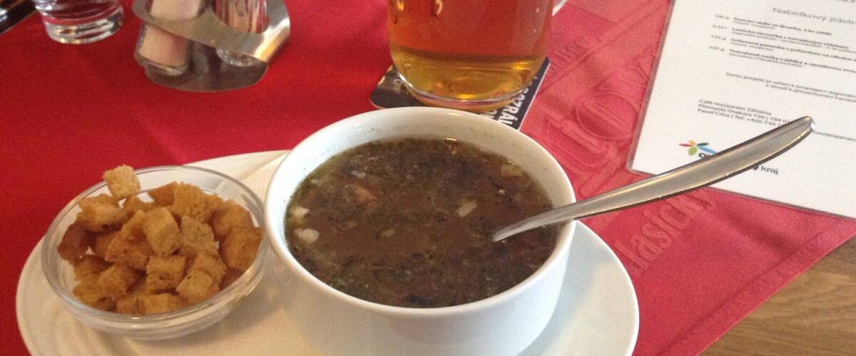 A bowl of soup with a spoon, a small bowl of croutons, and a glass of beer on a red tablecloth.