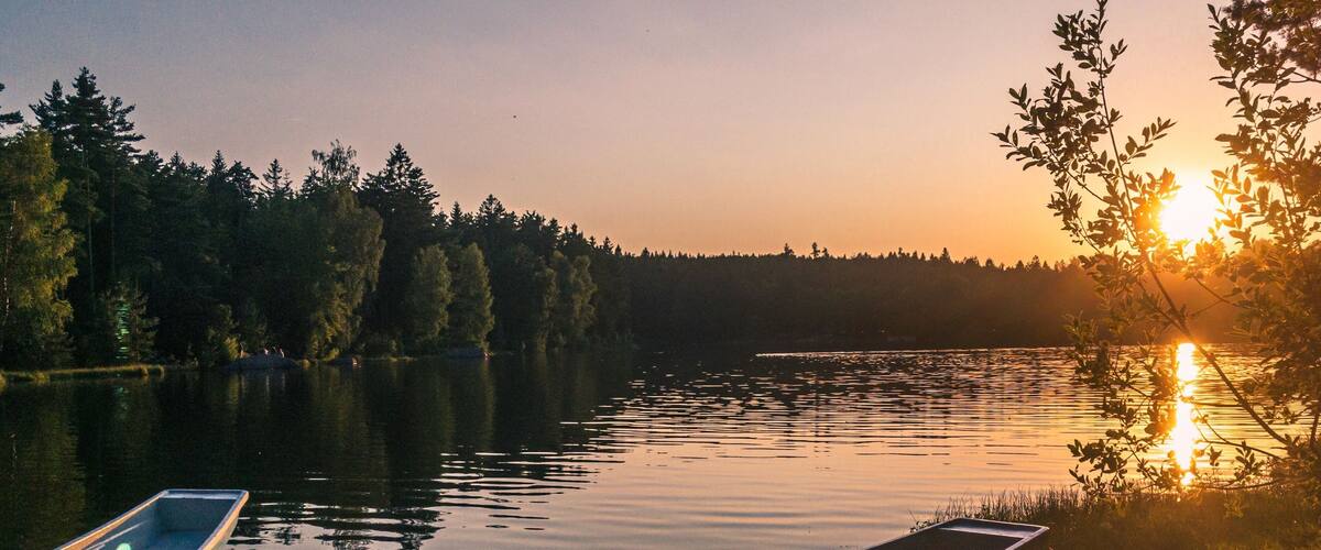 A sunset over a calm lake with boats docked on the shore and a forest in the background.