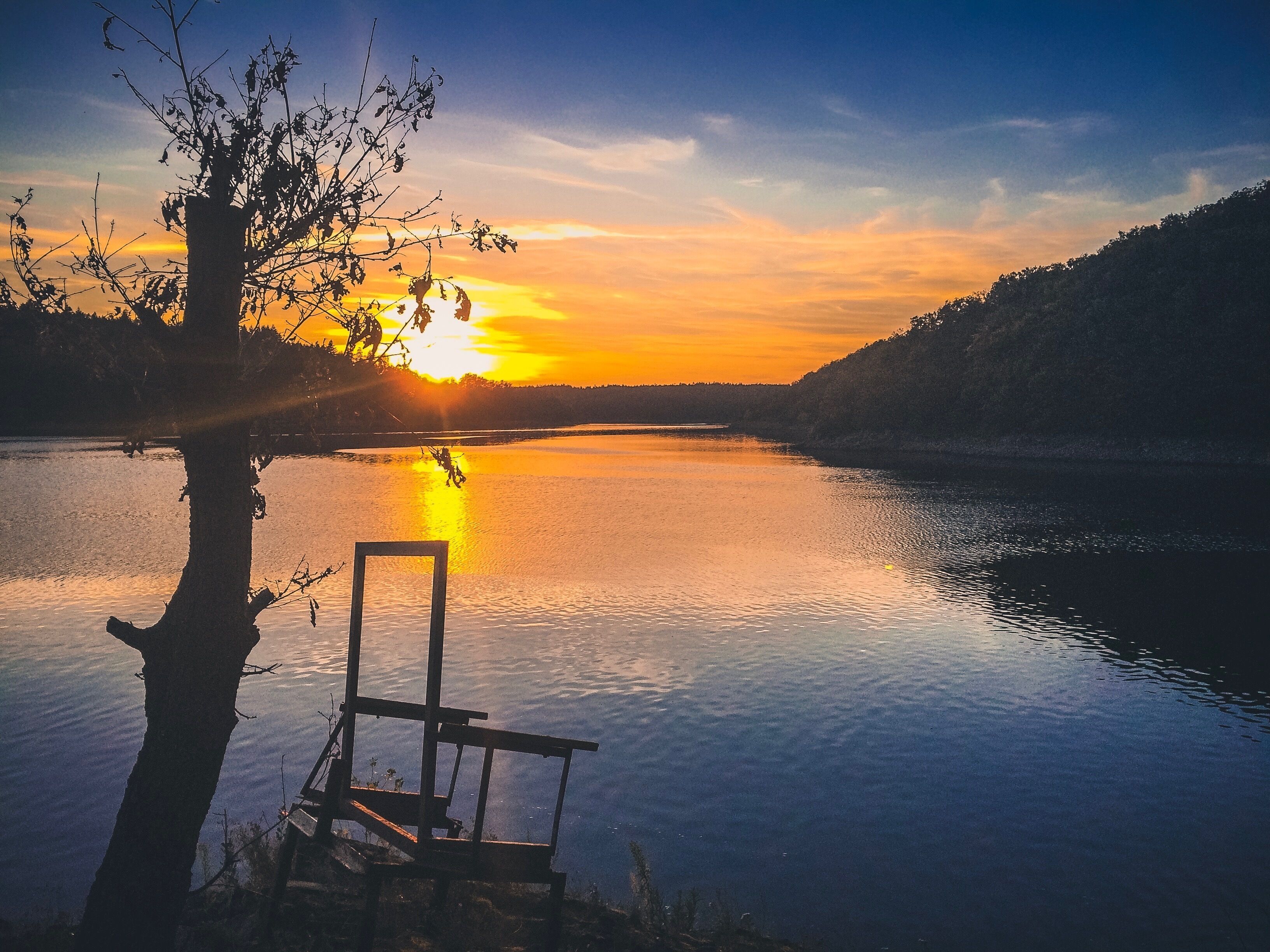 A sunset over a calm lake with a tree and a bench in the foreground.