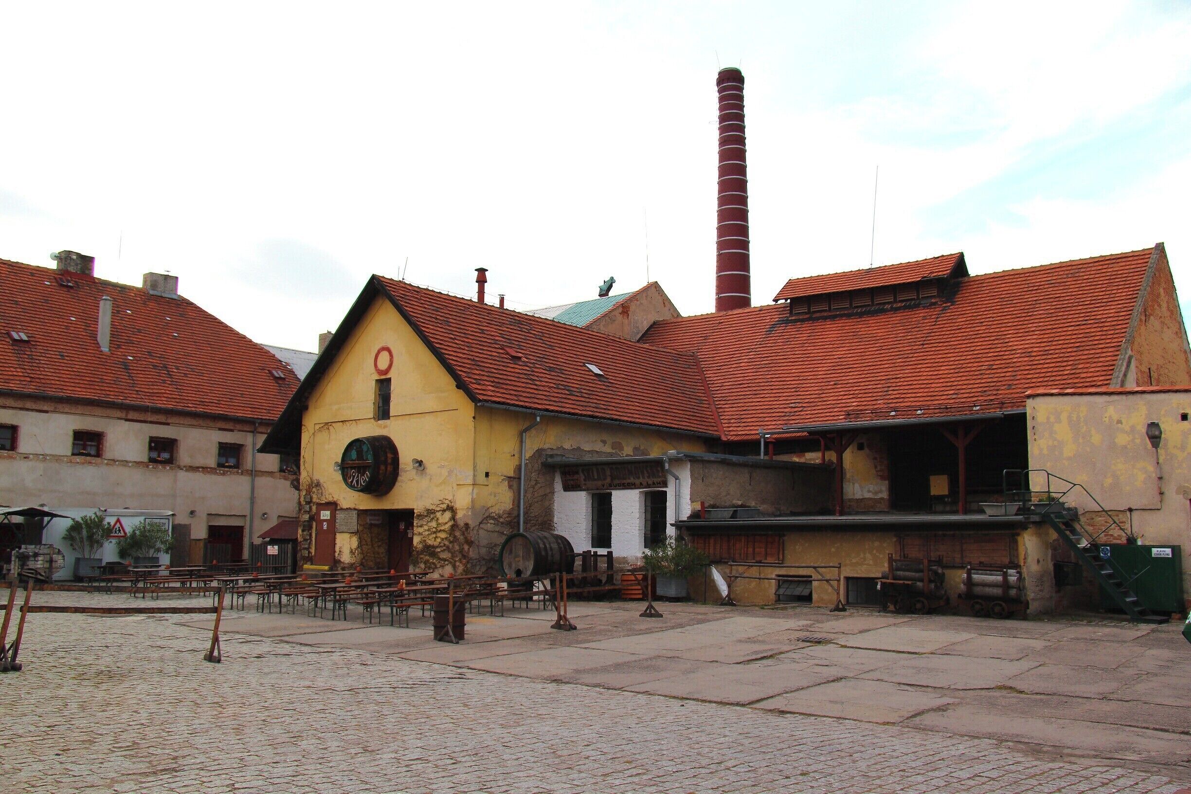 A cobblestone square with a yellow building, a red-roofed structure, and a tall chimney.