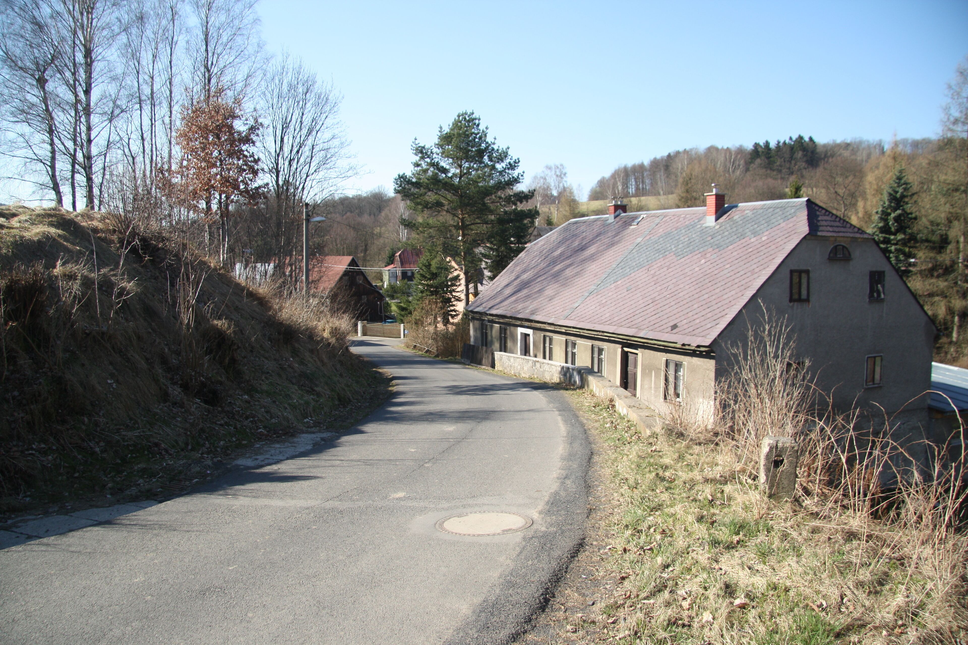 A rural road curving through a landscape with a stone house on the right and a hill on the left.