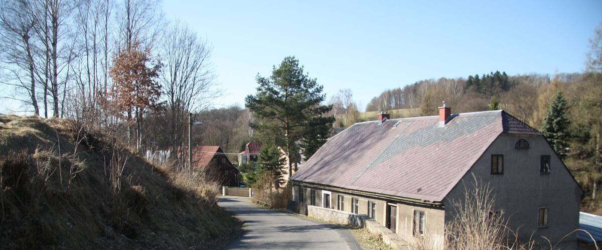 A rural road curving through a landscape with a stone house on the right and a hill on the left.