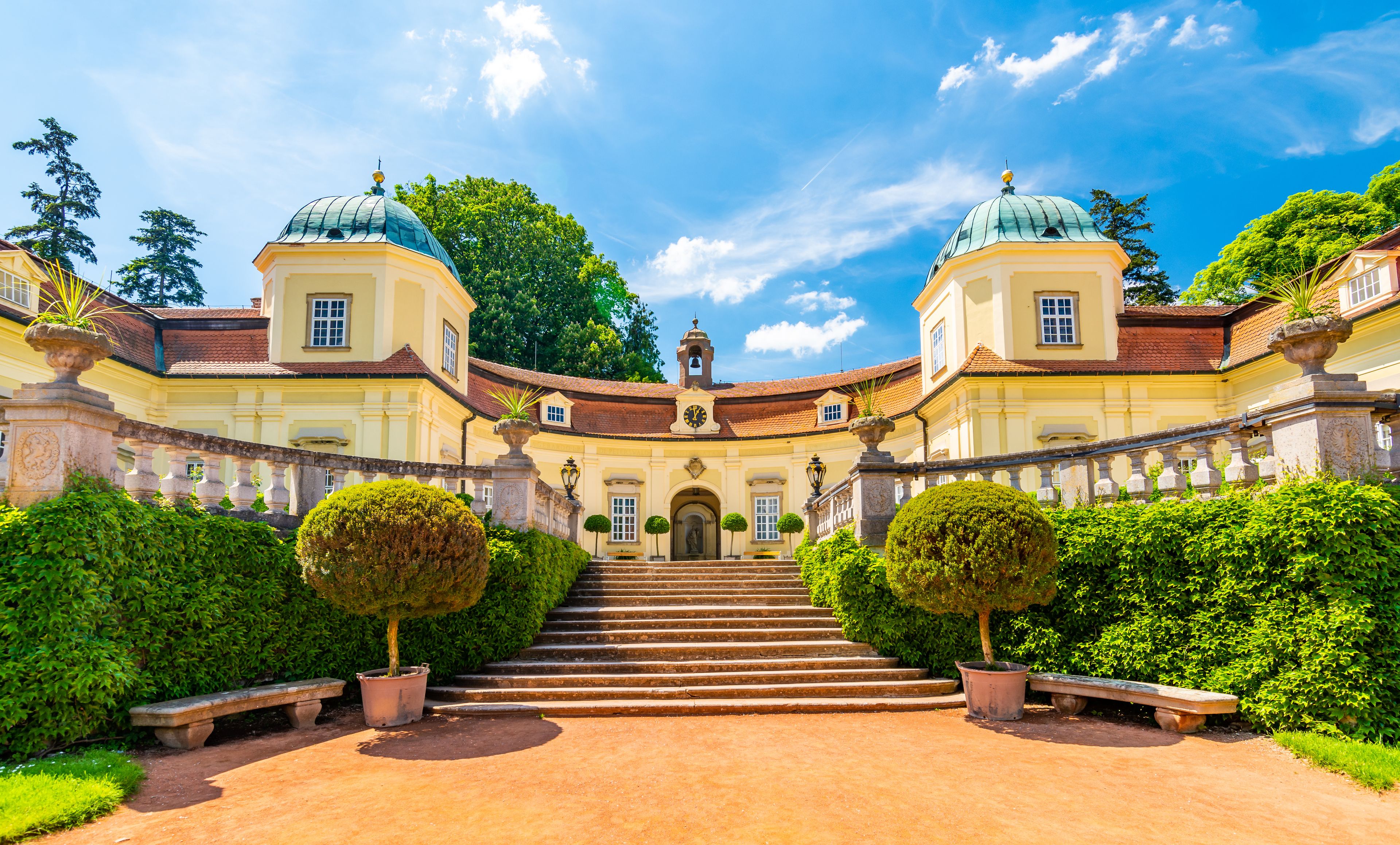 A grand building with two domed towers, a central staircase, and lush greenery.