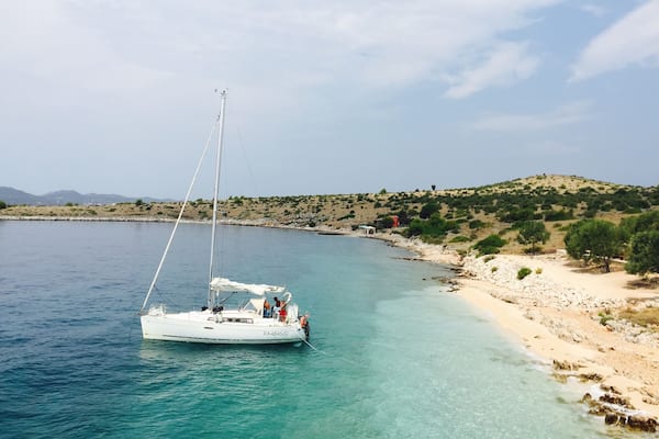 A sailboat on a calm sea near a rocky shoreline with sparse vegetation and a distant hill.
