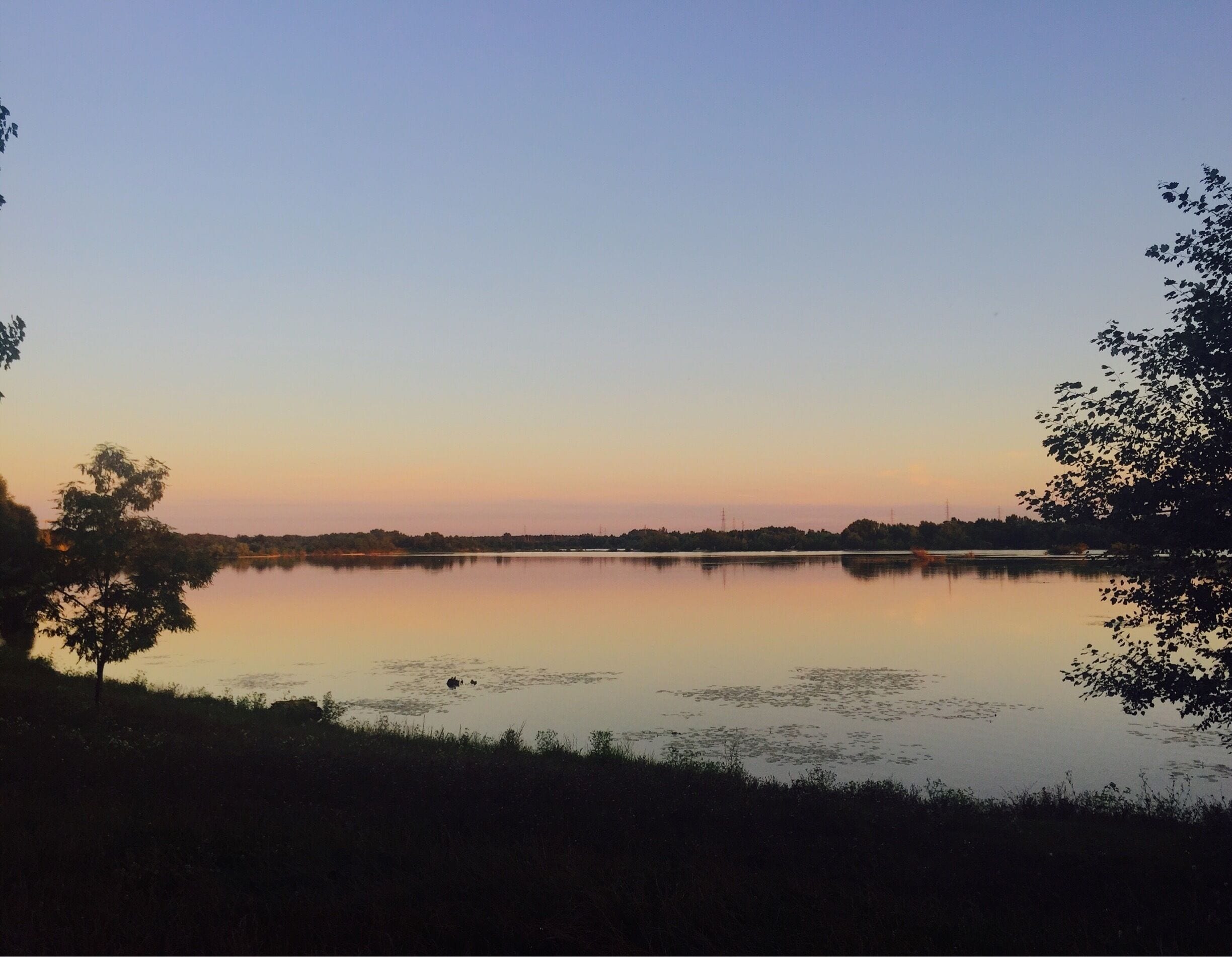 A calm lake at sunset with trees on the banks and a clear sky.