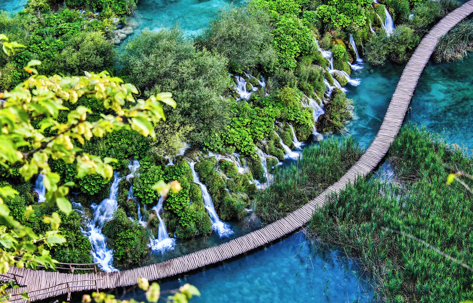 A wooden bridge over a clear blue river surrounded by lush greenery and cascading waterfalls.