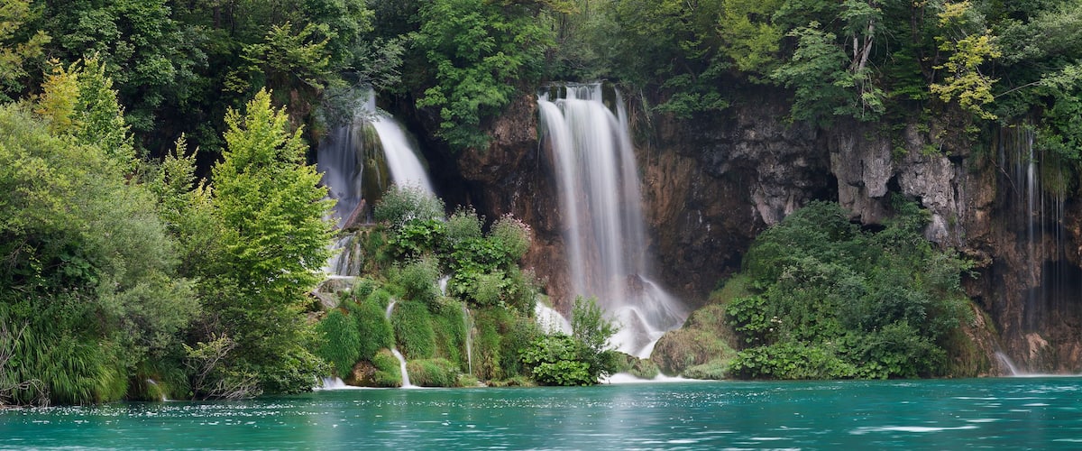 Una cascada se precipita en un lago turquesa rodeado de un bosque verde denso.
