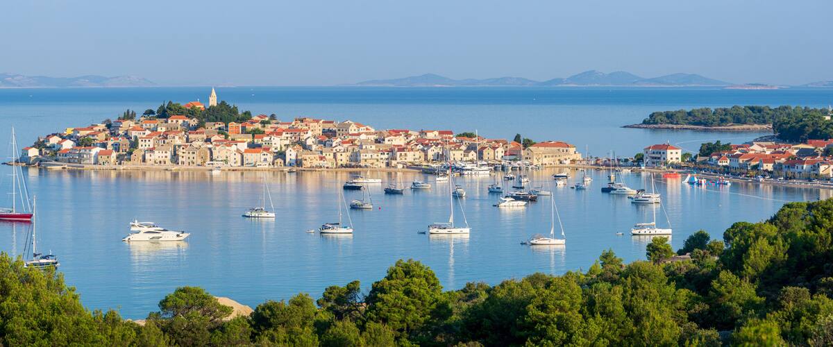 A coastal town with a dense cluster of buildings, a prominent church tower, and numerous boats docked in the harbor.