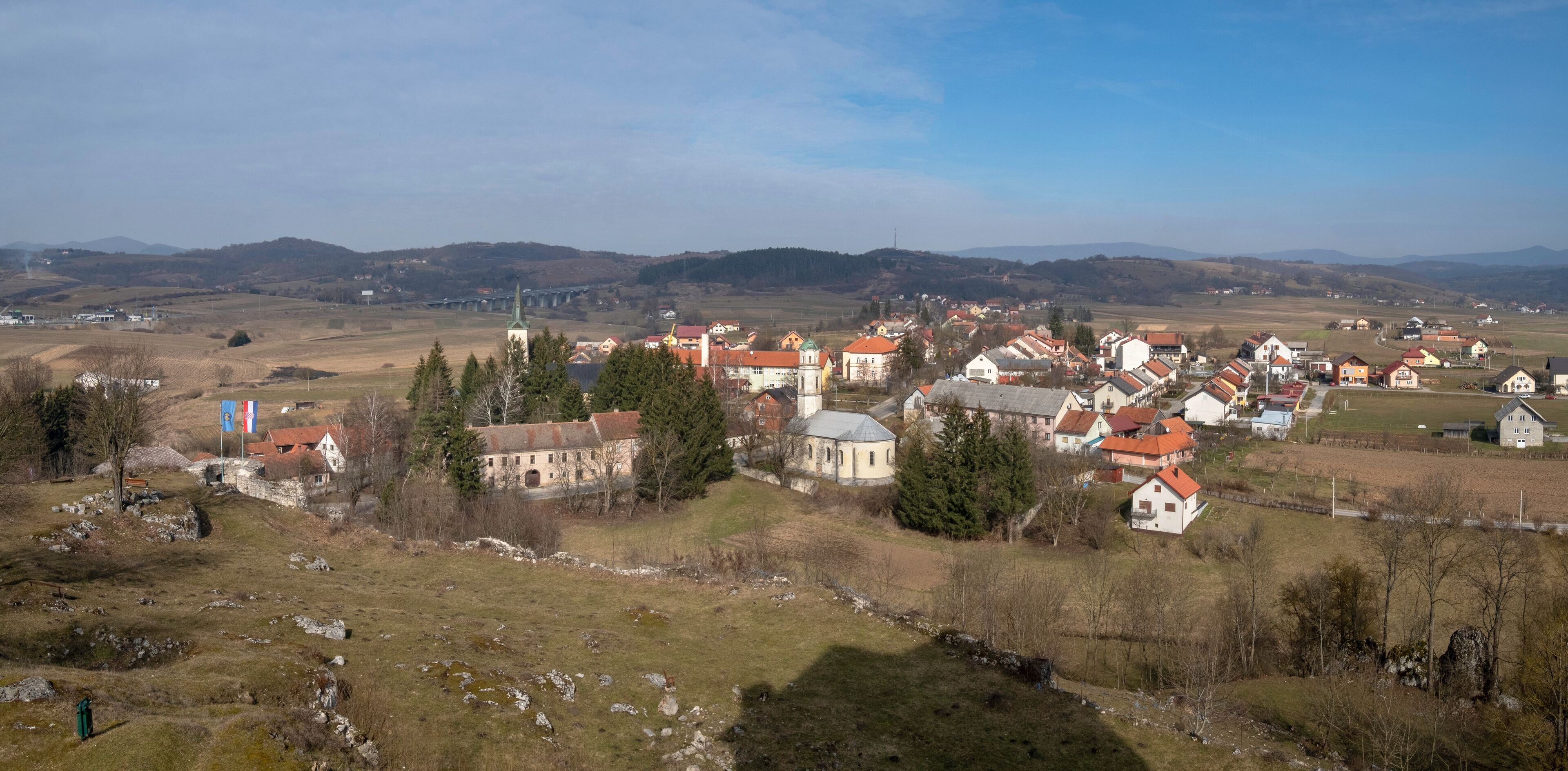 A village with red-roofed houses, a church, and a bridge in the background.