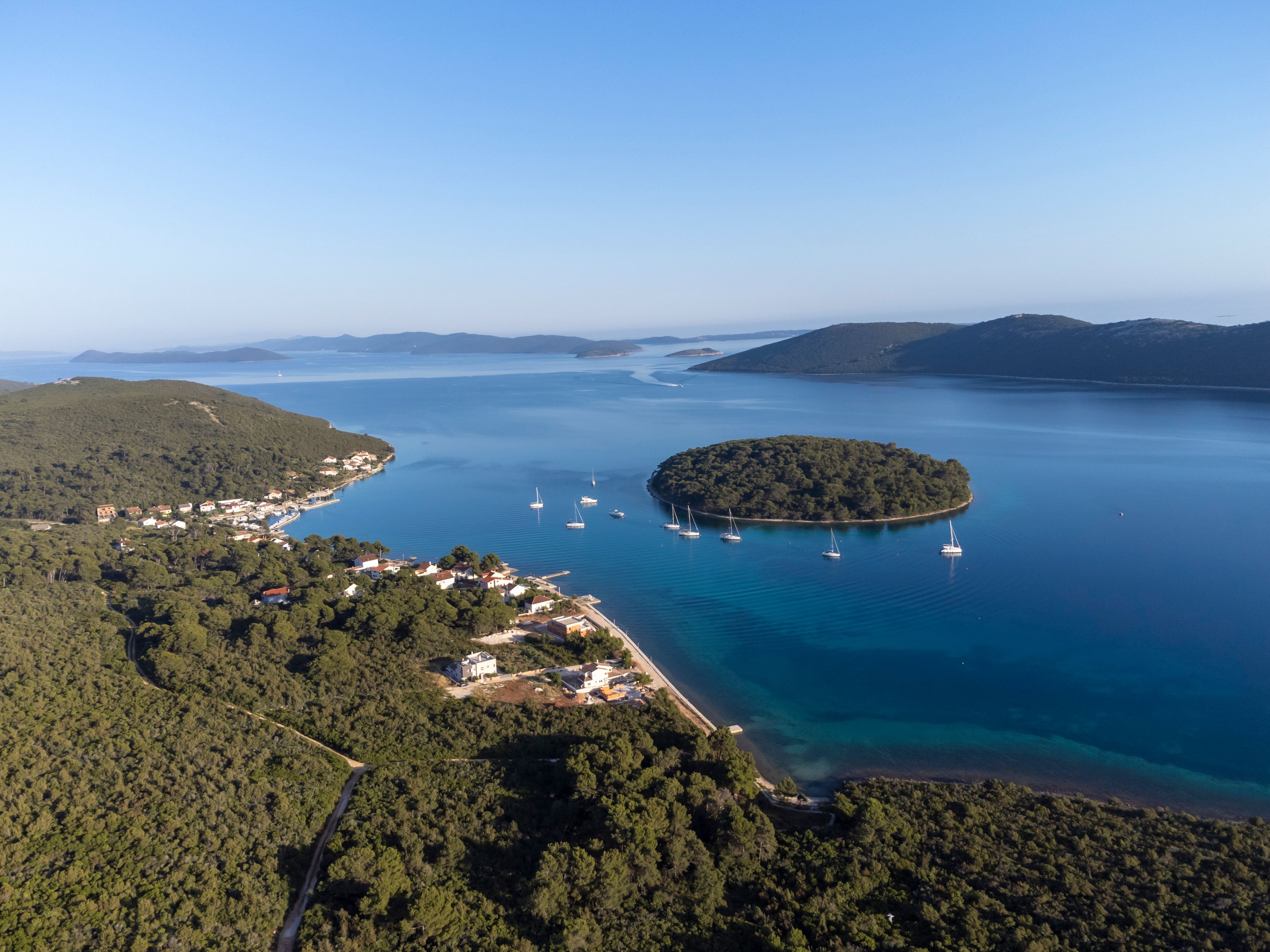 A coastal landscape with a small island surrounded by clear blue waters, a few sailboats, and a town with buildings and roads.