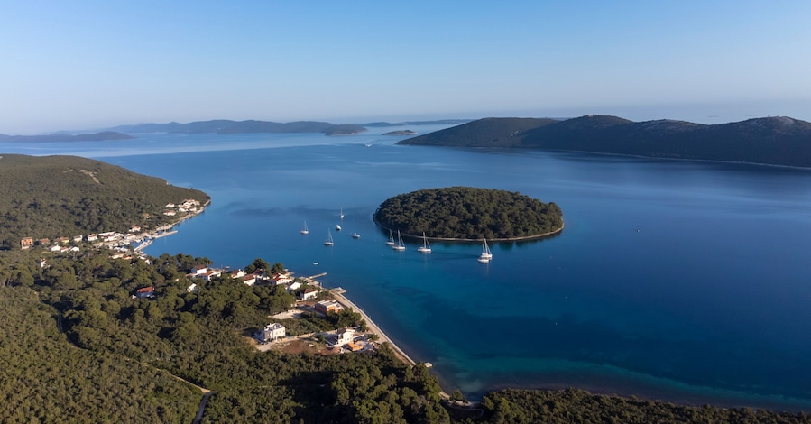 An aerial view of a coastal town with a small island surrounded by clear blue waters, several sailboats anchored near the shore, and a backdrop of distant hills.