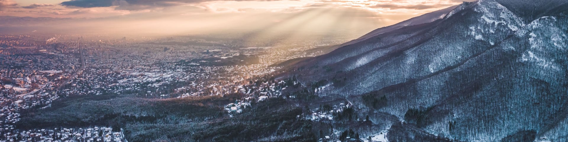 A mountainous landscape with a city in the valley below, a prominent peak with a tower, and rays of sunlight piercing through the clouds.