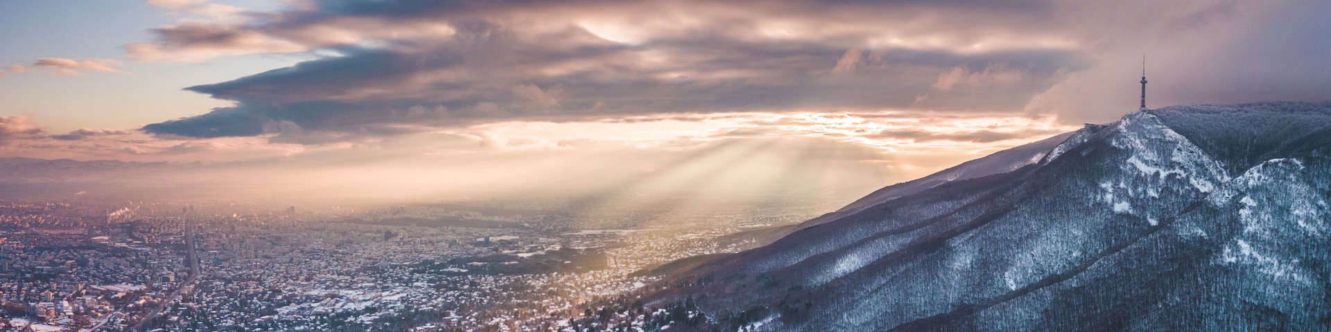 A mountainous landscape with a city in the valley below, a prominent peak with a tower, and rays of sunlight piercing through the clouds.