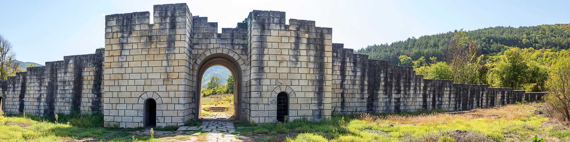 Un arco de piedra que da paso a una zona boscosa con una montaña al fondo.