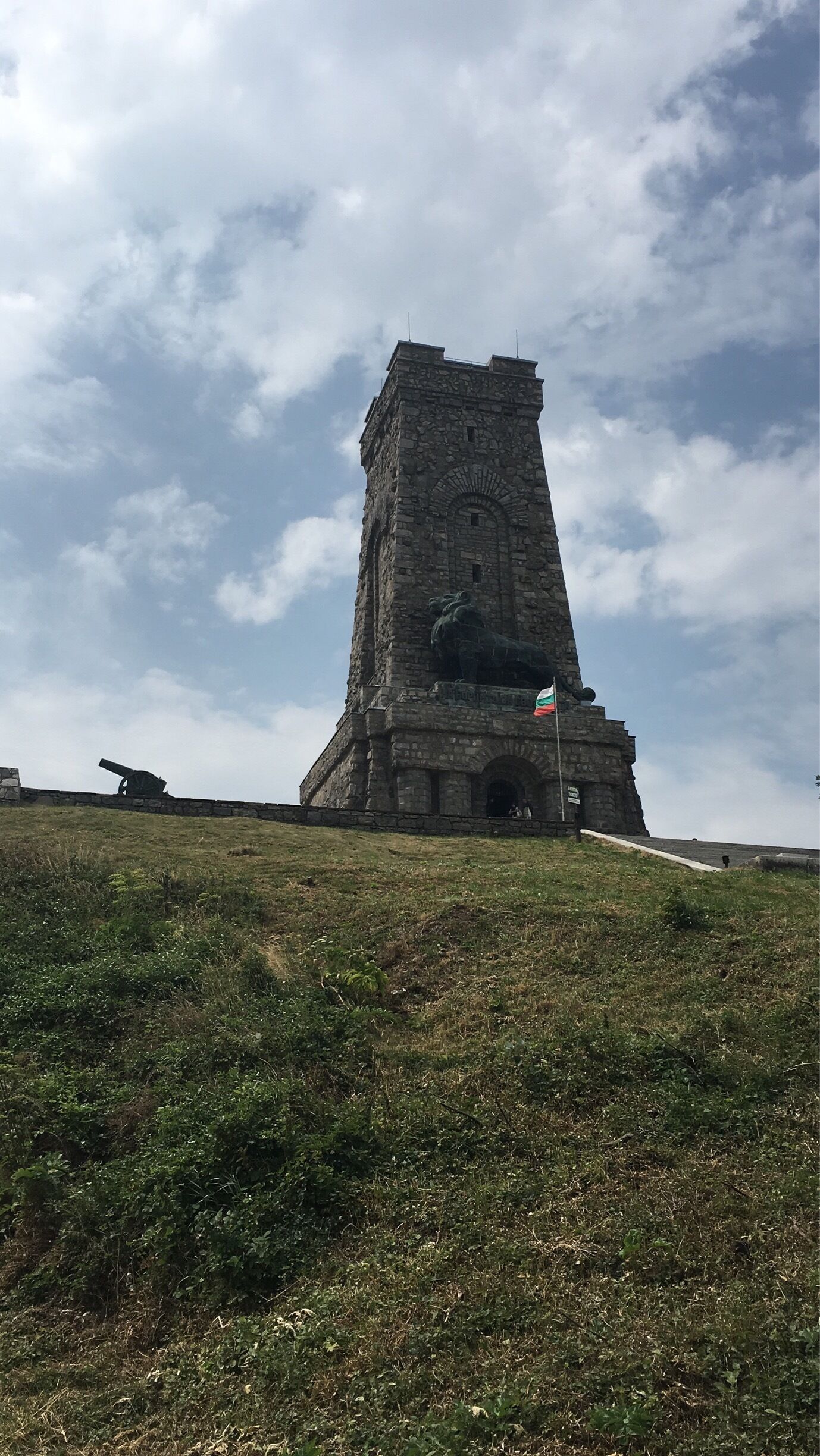 A tall stone tower with a flag on top, surrounded by grass and a cloudy sky.