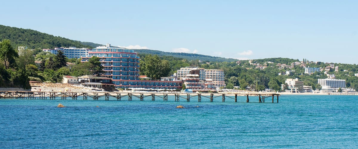 A coastal cityscape with a pier extending into the sea, surrounded by lush green hills and various buildings.