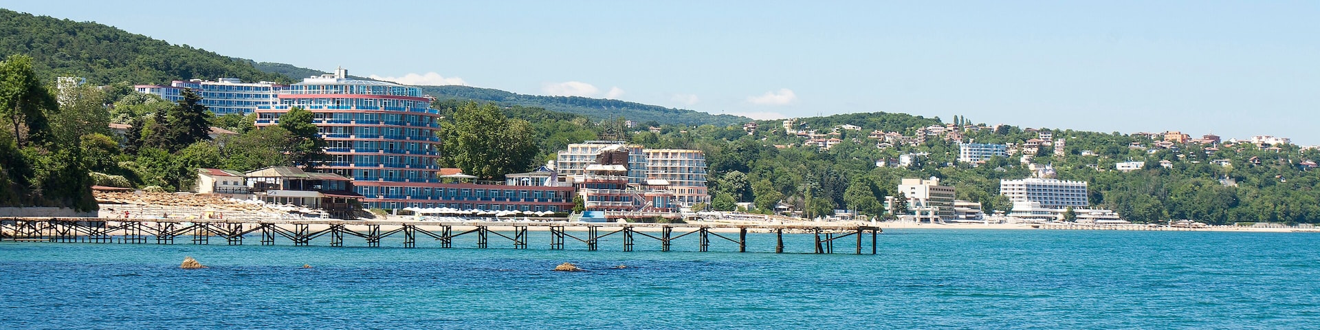 A coastal cityscape with a pier extending into the sea, surrounded by lush green hills and various buildings.