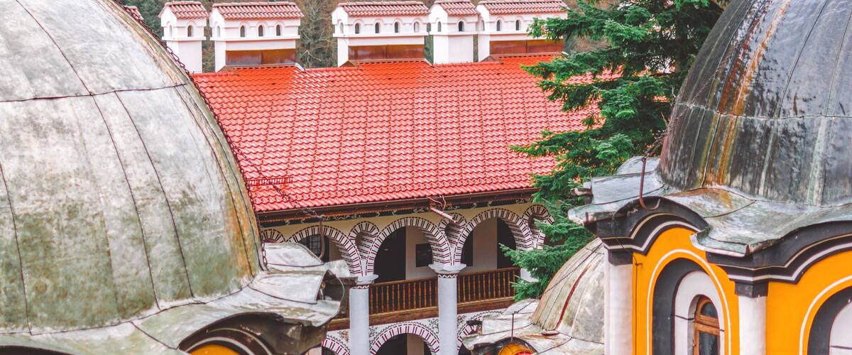 A historic building with domed architecture and a red-tiled roof, surrounded by a forested hillside.
