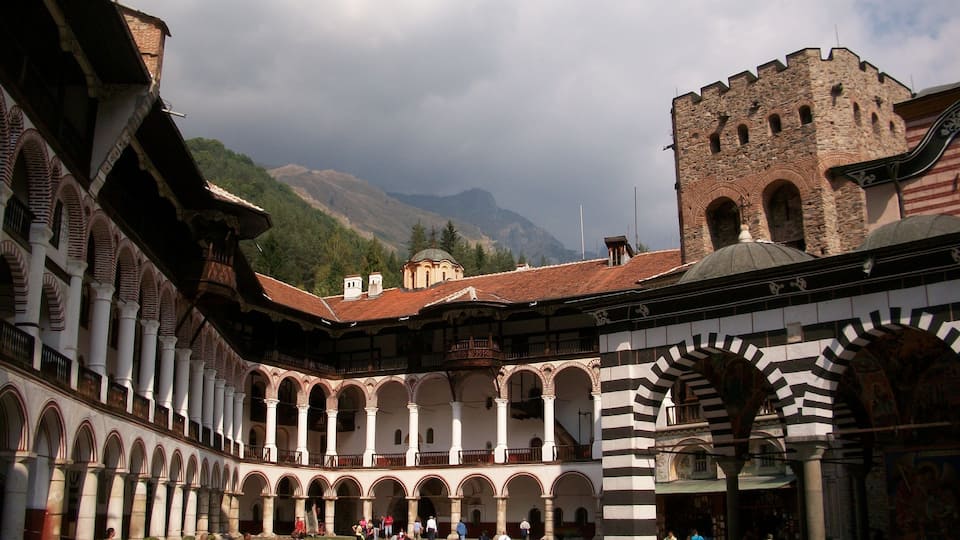 A historic courtyard with arches, a tower, and mountains in the background.