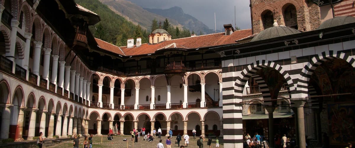 A historic courtyard with arches, a tower, and mountains in the background.