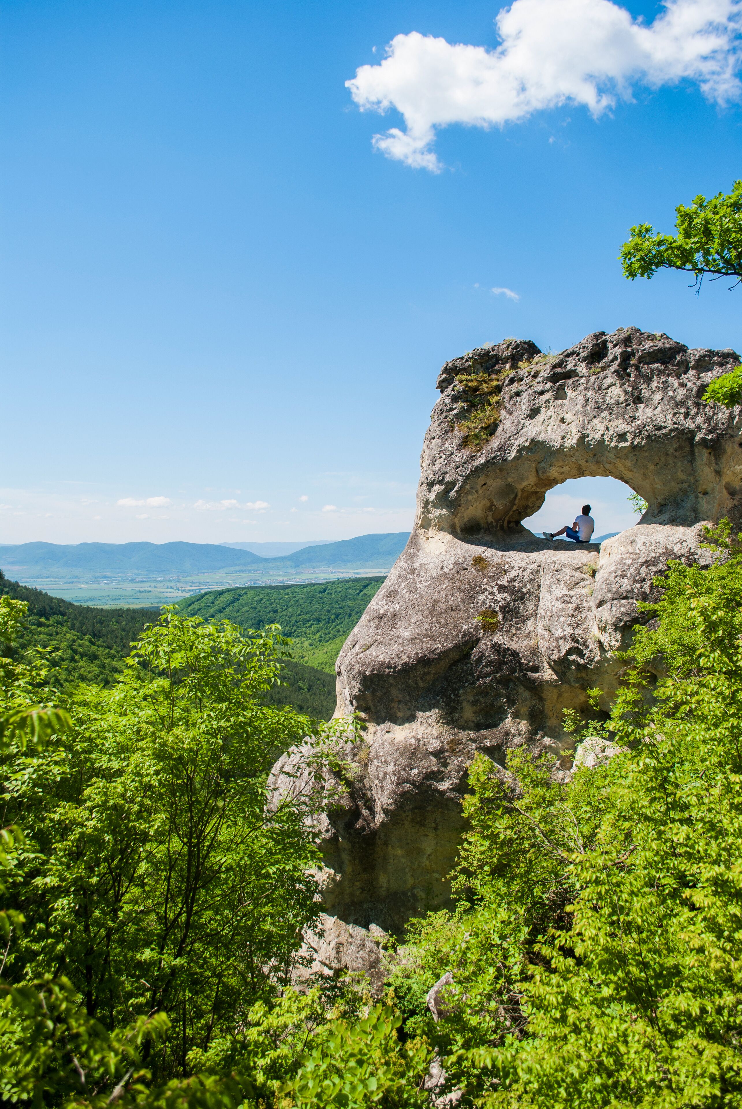 A person sitting in a natural rock arch with a scenic view of a valley and mountains in the background.