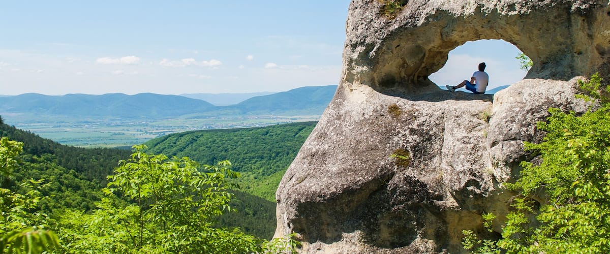 A person sitting in a natural rock arch with a scenic view of a valley and mountains in the background.